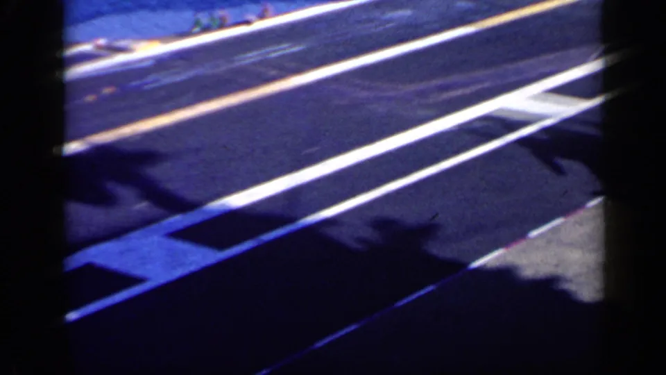 Final 8mm frame showing painted aircraft carrier deck lines near blue water, with shadows and only partial aircraft visible.