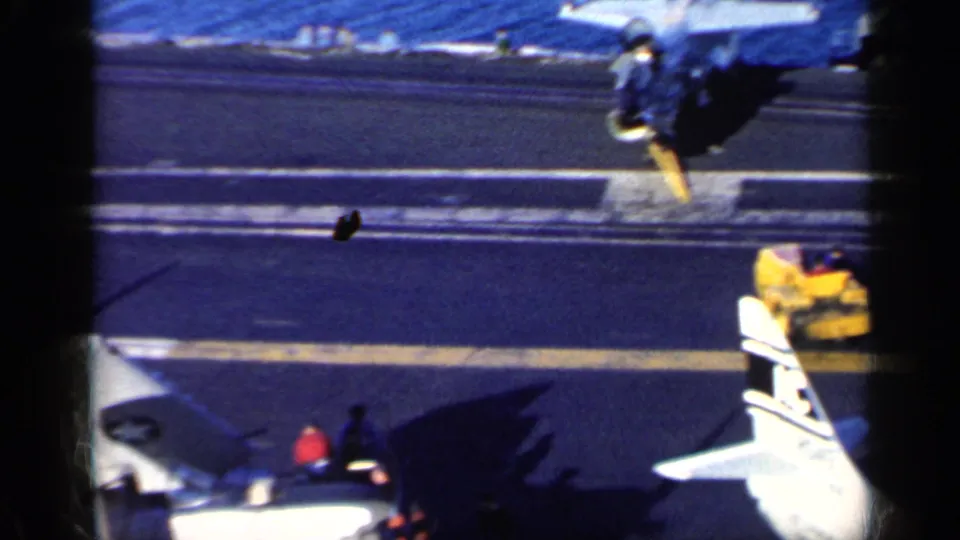 1962 8mm frame showing military aircraft on a carrier deck near Cuba, with U.S. insignia, deck markings, and ocean beyond.
