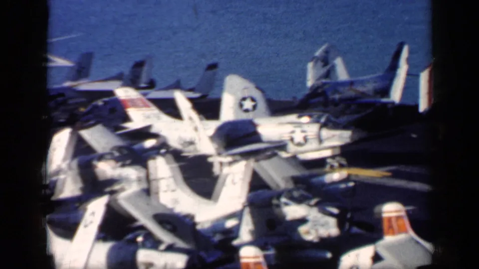 1962 8mm frame from near Cuba showing a crowded U.S. carrier deck with multiple aircraft, folded wings, and insignia.
