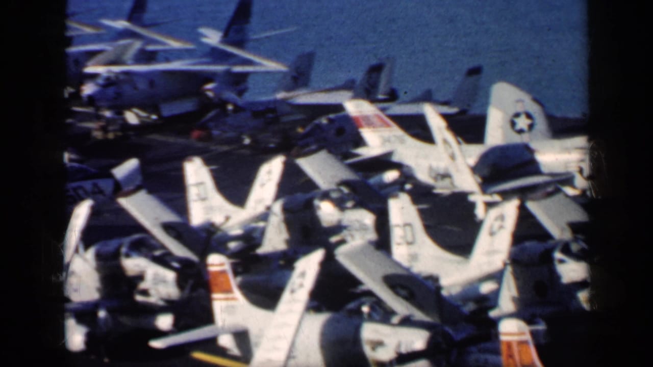 Rows of U.S. Navy jets parked on an aircraft carrier deck beside blue ocean.