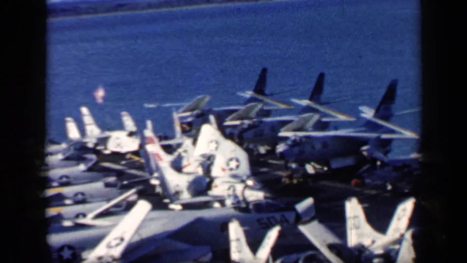 1962 Cuba-context carrier-deck view with multiple U.S. Navy jets, visible insignia, folded wings, and deep blue water beyond the ship.