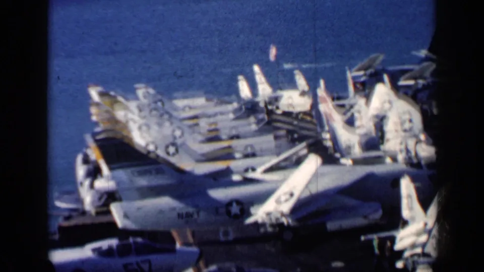 1962 Cuba-context 8mm frame showing carrier-deck aircraft lined up near the deck edge, with Navy markings and ocean visible.