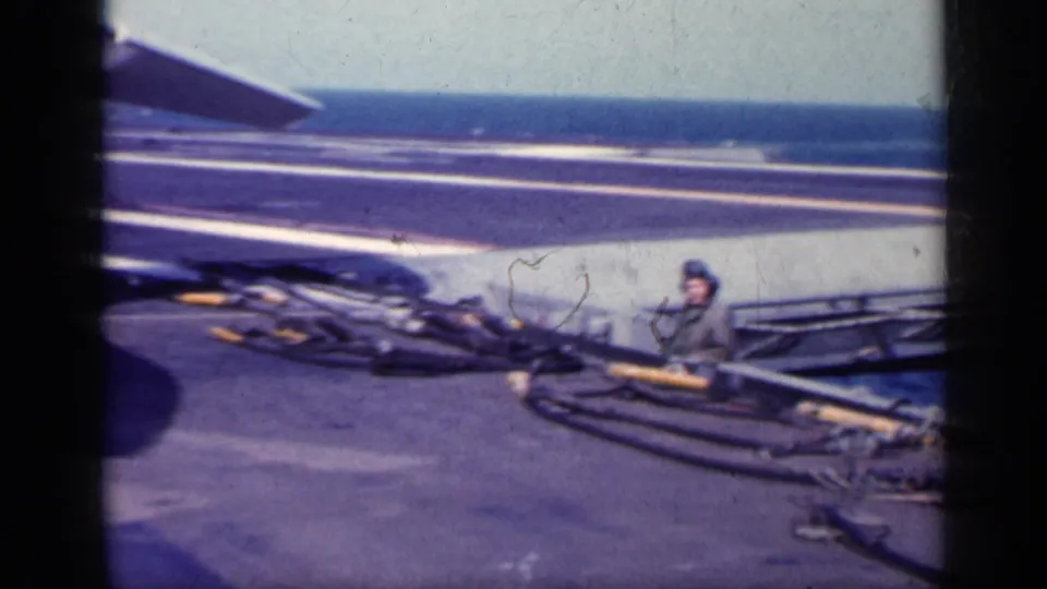 1962 Cuba-associated 8mm frame showing a helmeted sailor near coiled deck cables on an aircraft carrier with the ocean beyond.