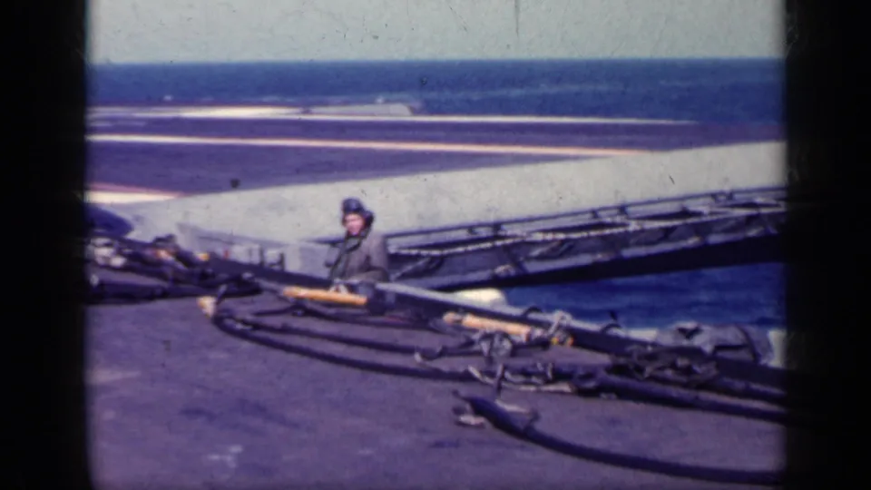 Helmeted sailor on an aircraft carrier deck near hoses or cables, with open sea and film-frame borders visible.