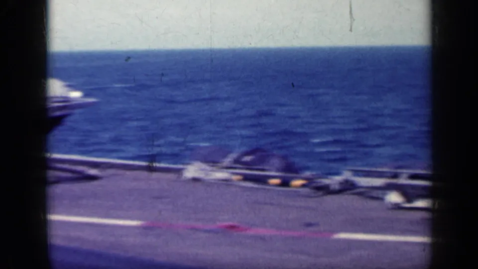 Carrier deck edge with scattered equipment and markings, framed by open sea in grainy 8mm footage.