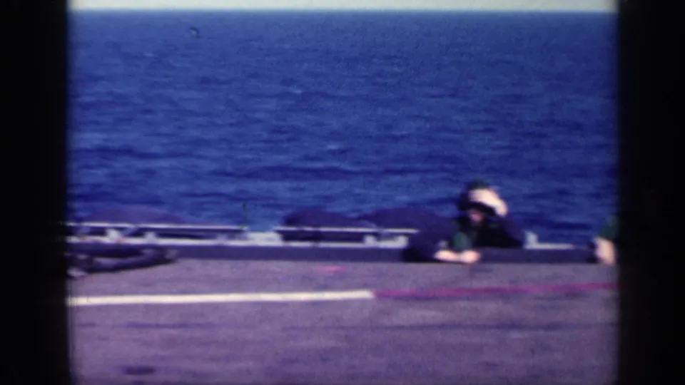 Helmeted sailor crouched near the carrier deck edge with open water behind, seen in faded 8mm color.