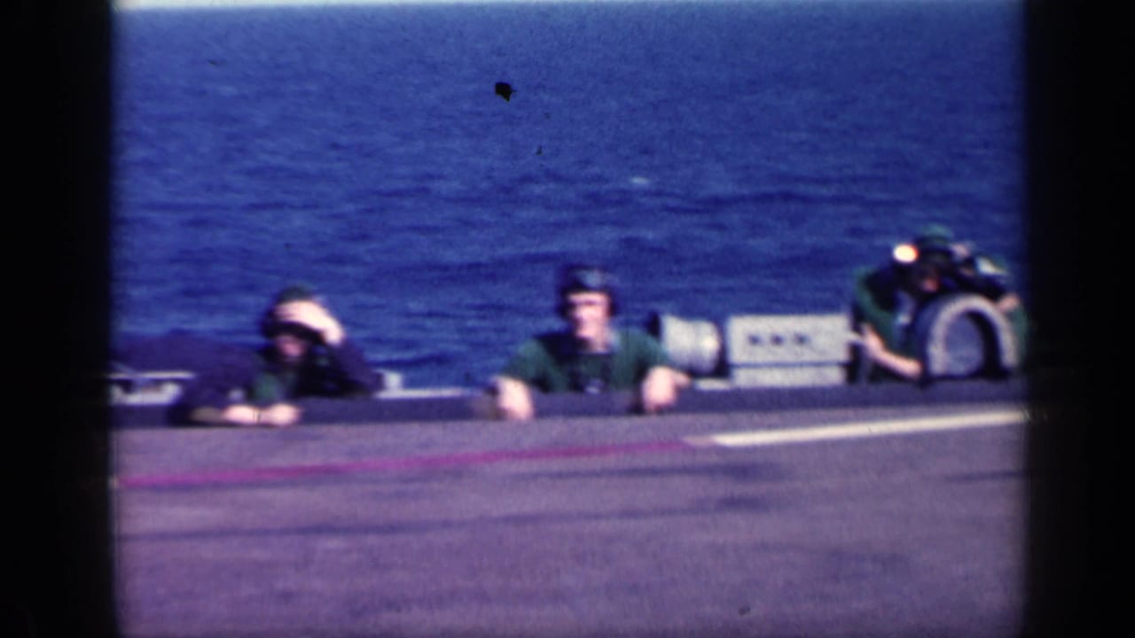 Two helmeted crew members visible behind an aircraft or deck edge with ocean in the background.
