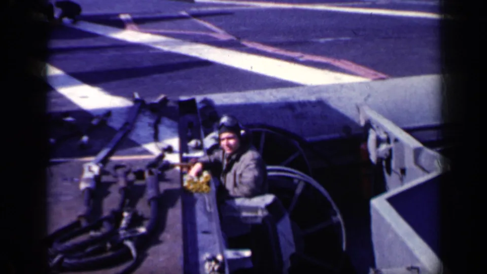 A crew member in helmet and work clothing stands beside aircraft carrier deck equipment and painted markings in the 1962 Cuba-linked footage.