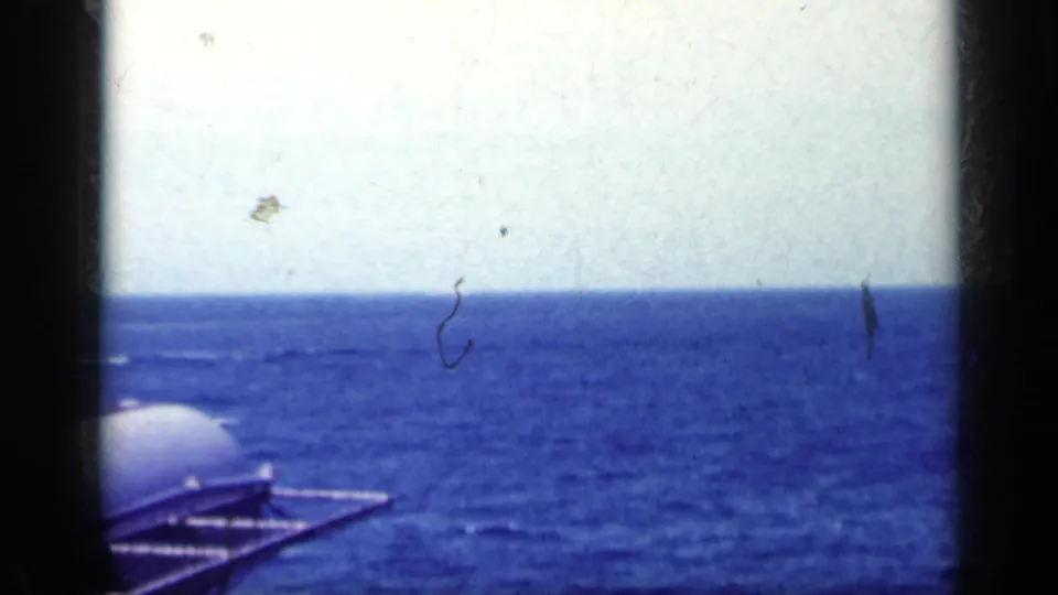 Open sea and horizon fill the frame, with a curved aircraft or ship component entering at lower left in grainy 8mm footage.