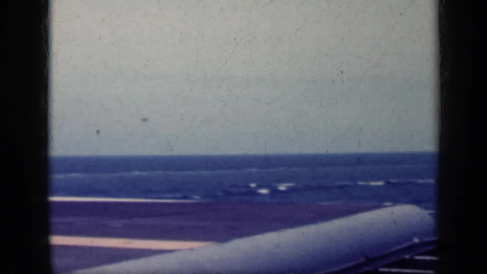 The carrier deck and ocean horizon are seen beyond a large aircraft surface or ship structure in the 1962 Cuba-linked home-movie footage.