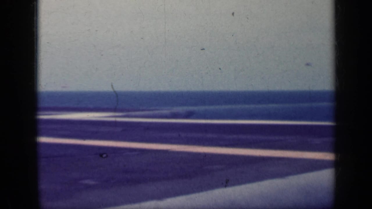 1962 Cuba-associated 8mm frame showing a carrier flight deck, white deck markings, ocean horizon, and dark foreground ship structure.