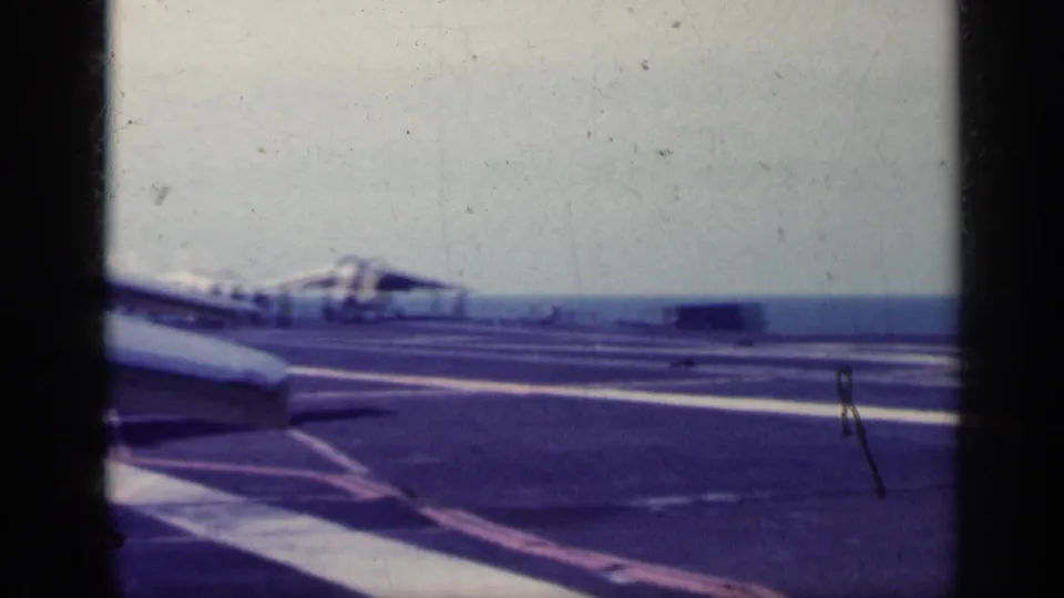 1962 Cuba-associated 8mm frame showing a jet on a marked aircraft carrier deck, with ocean and pale sky beyond.