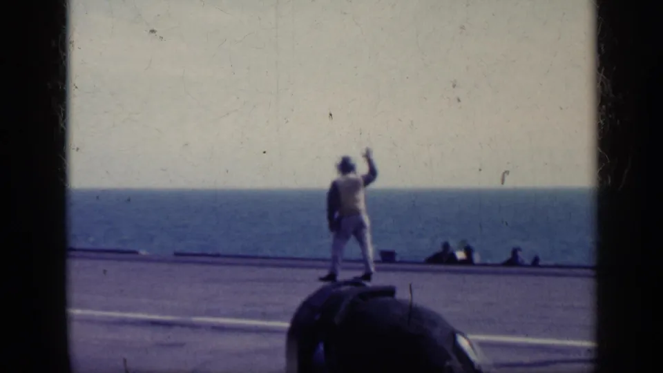 1962 Cuba-associated 8mm frame showing a light-clothed deck crew figure signaling on a carrier deck, with ocean in the background.
