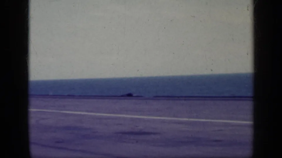 1962 Cuba-associated 8mm frame showing an open section of carrier deck, white markings, ocean horizon, and pale sky.