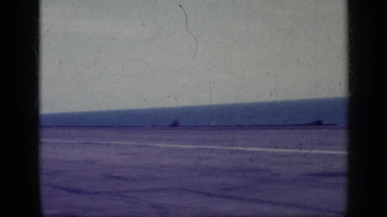 Wide tarmac beside an ocean horizon with film gate edges and no central figure visible.