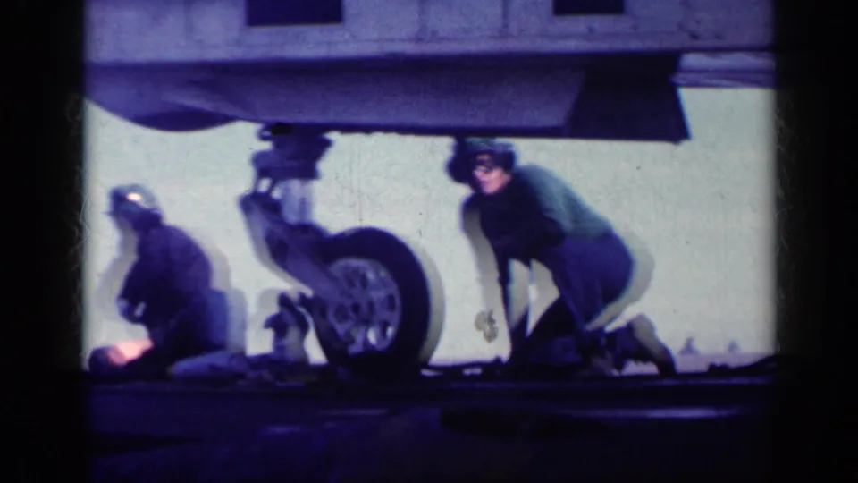 Military crew in deck clothing work under a U.S. Navy aircraft landing gear in metadata-supported 1962 Cuba footage.