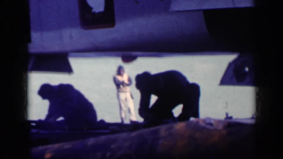 Carrier deck personnel work beneath the aircraft underside while a uniformed figure stands in the background in this 1962 Cuba frame.