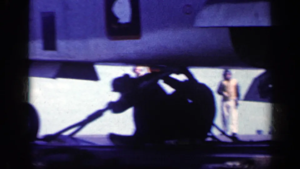 Dark silhouettes of crew handle equipment under an aircraft on a bright carrier deck, with another person standing in the background.