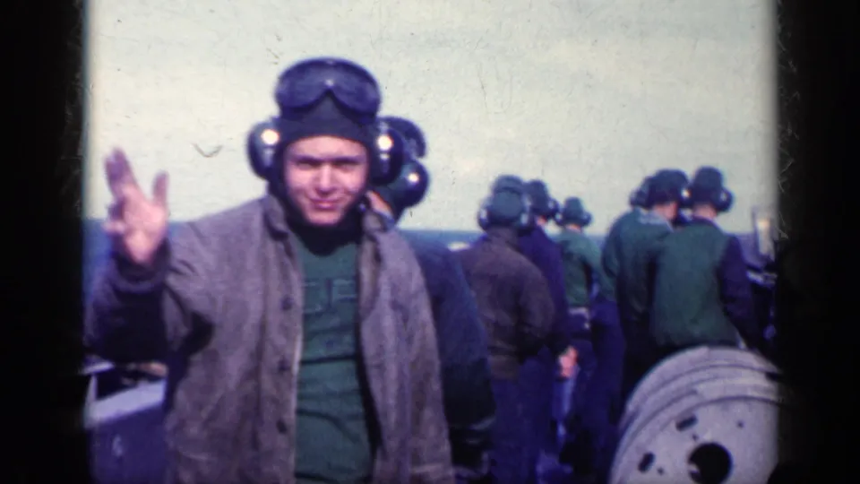 A man in vintage flight gear waves toward the camera while other helmeted personnel gather on the carrier deck in the background.