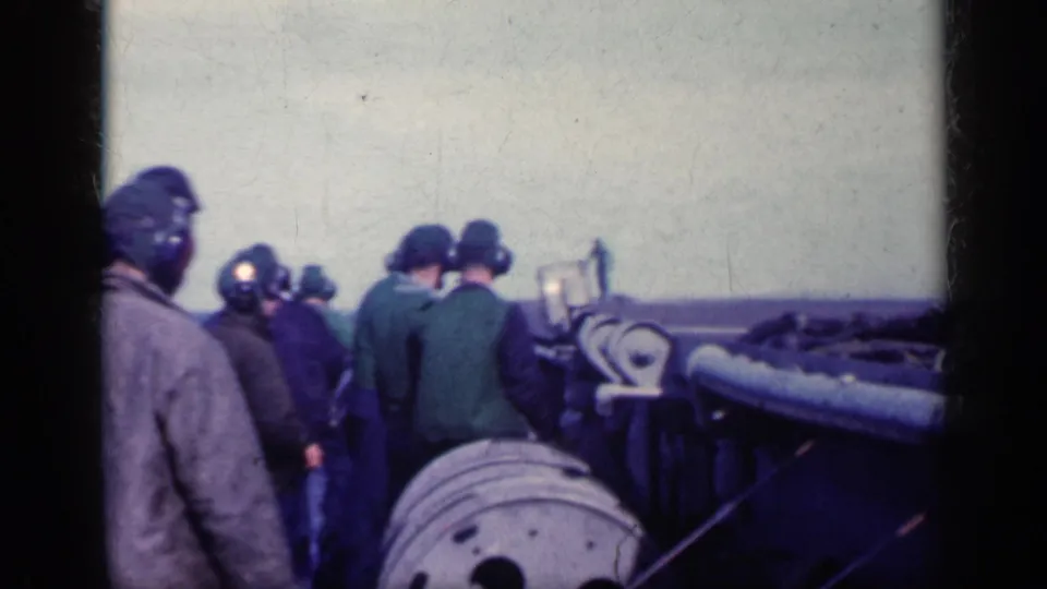 Several helmeted crew members stand near aircraft parts or deck gear on a carrier, with open water visible along the horizon.