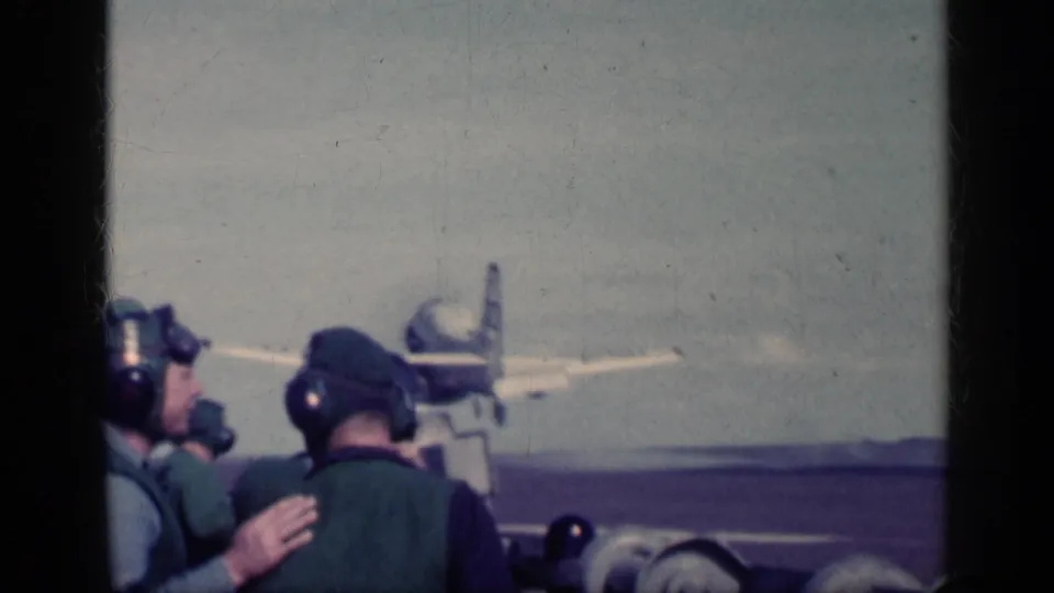 U.S. Navy deck personnel in headsets watch an aircraft ahead on the flight deck, with sea or distant land at the horizon.