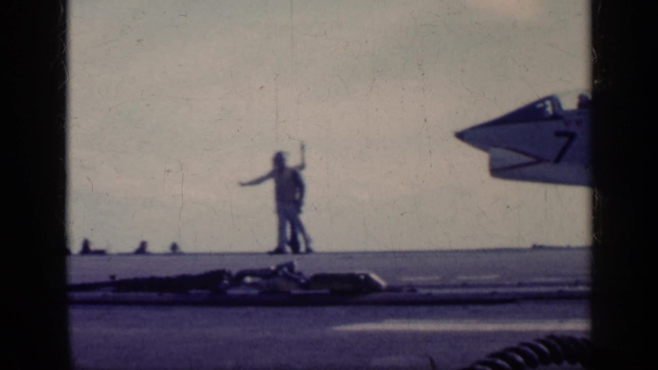 Deck crew signal near the nose of a naval jet on an aircraft carrier.