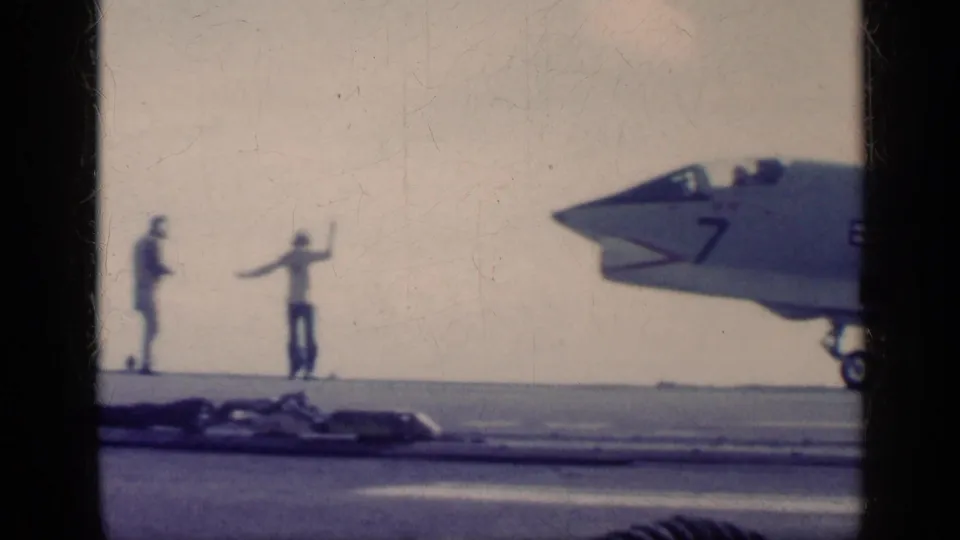 1962 Cuba-era 8mm frame with two deck crew signaling while a marked naval jet sits at the right edge of the carrier deck.