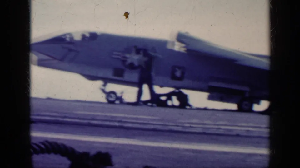 1962 Cuba-era 8mm frame showing the side of a U.S. Navy jet with star insignia and deck crew nearby on a carrier.