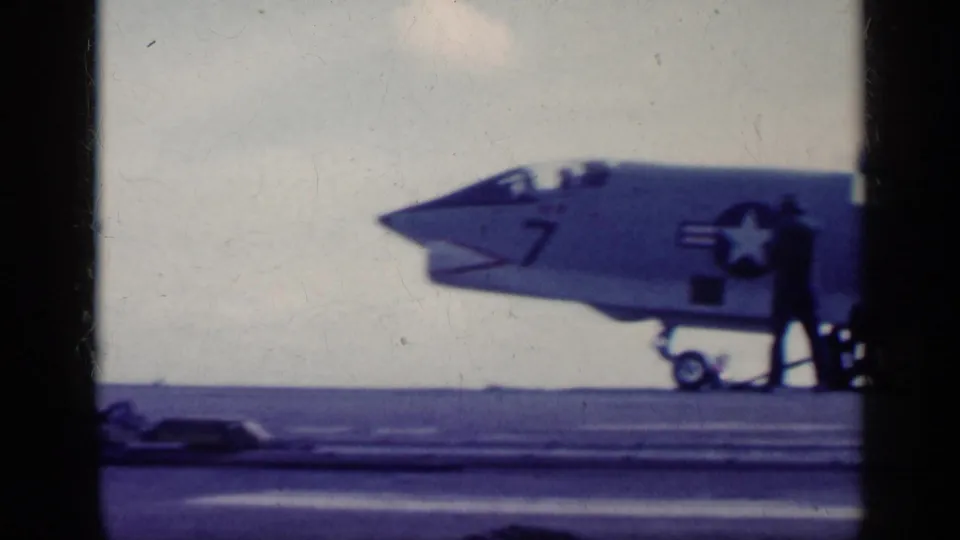 1962 Cuba-era 8mm frame of deck crew beside the nose and landing gear of a U.S. Navy jet marked 7 on a carrier deck.