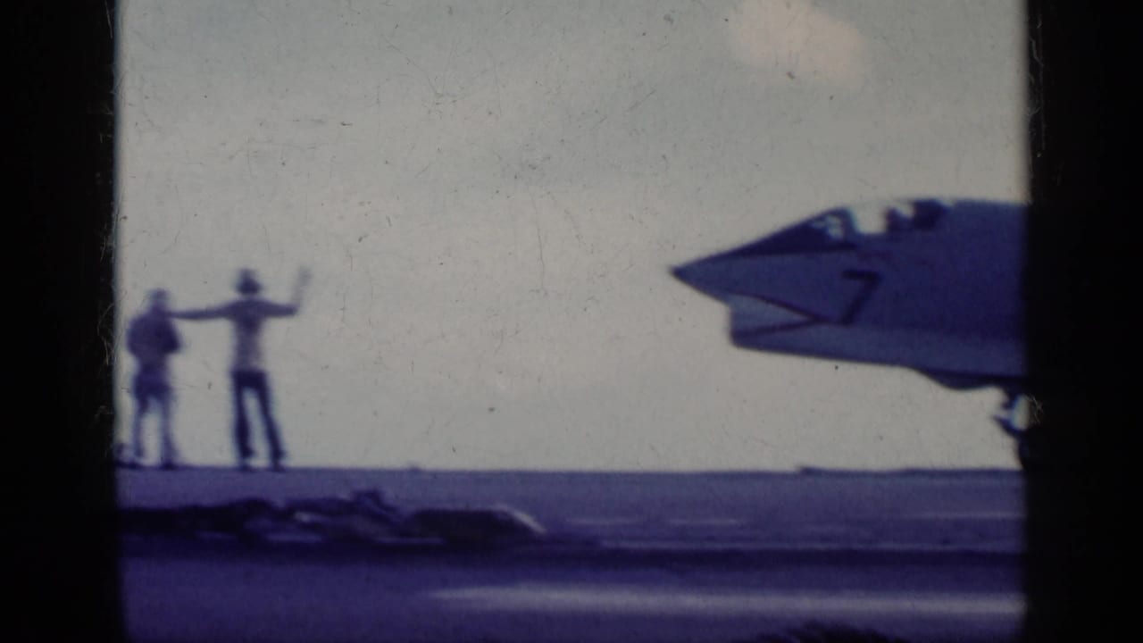 Two deck crew members signal beside a military jet nose on an aircraft carrier deck.