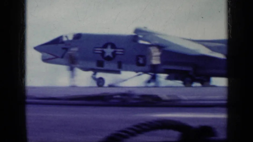 1962 8mm frame showing a U.S. Navy jet marked number 7 on an aircraft carrier deck, with insignia and landing gear visible.