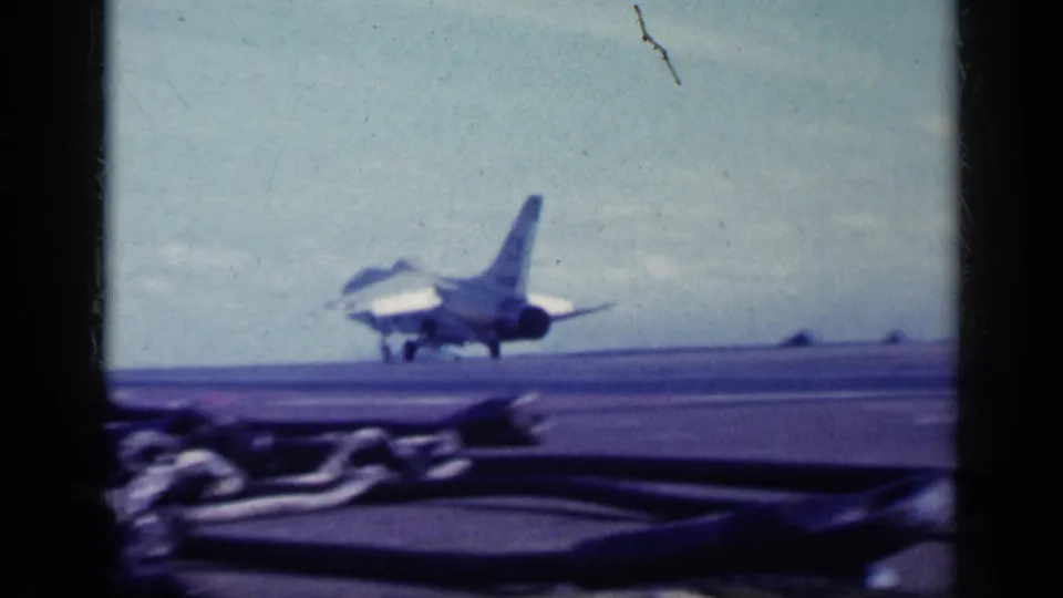 1962 8mm frame showing a rear view of a naval jet on the carrier deck, with foreground deck gear and open sky beyond.