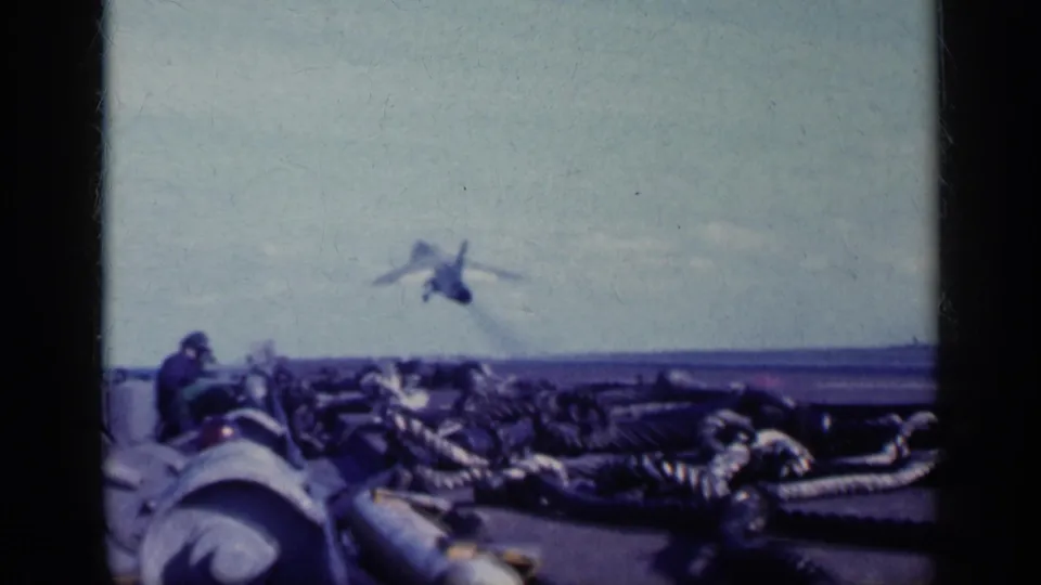 1962 8mm frame near Cuba showing a jet airborne beyond the carrier deck, with coiled deck gear and a crew member in the foreground.
