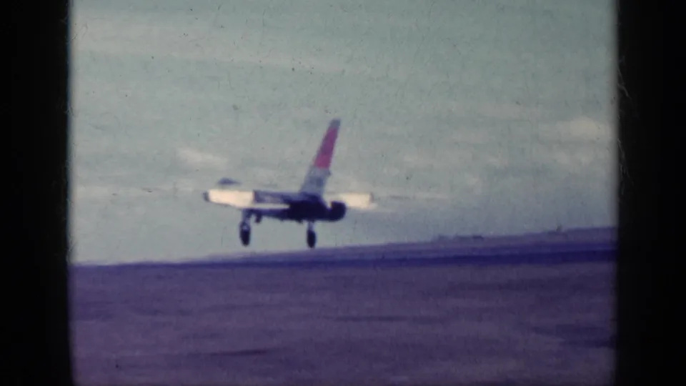 1962 Cuba-labeled 8mm frame showing a military jet airborne above the carrier deck, with landing gear visible against a cloudy sky.