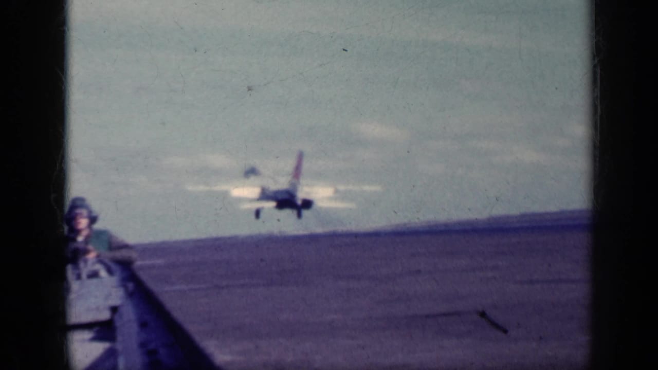 Military jet near the carrier deck with a helmeted crew member in the foreground.