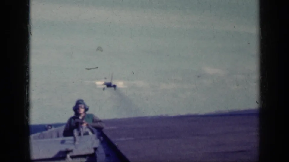 A helmeted crew member stands on the aircraft carrier deck as a military jet appears farther downrange against the ocean horizon.