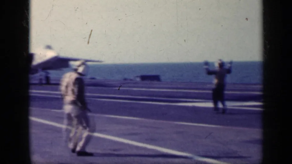 On the aircraft carrier deck, crew members stand near painted runway lines while a military jet is partly visible at the left of frame.