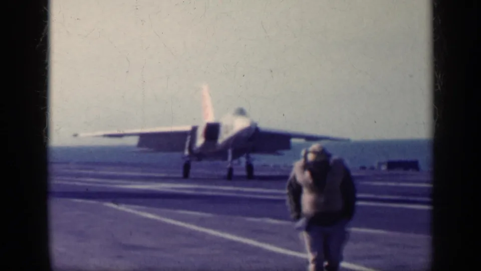A military jet sits or rolls on the carrier deck as a crew member bends in the foreground, with the ocean horizon behind the aircraft.