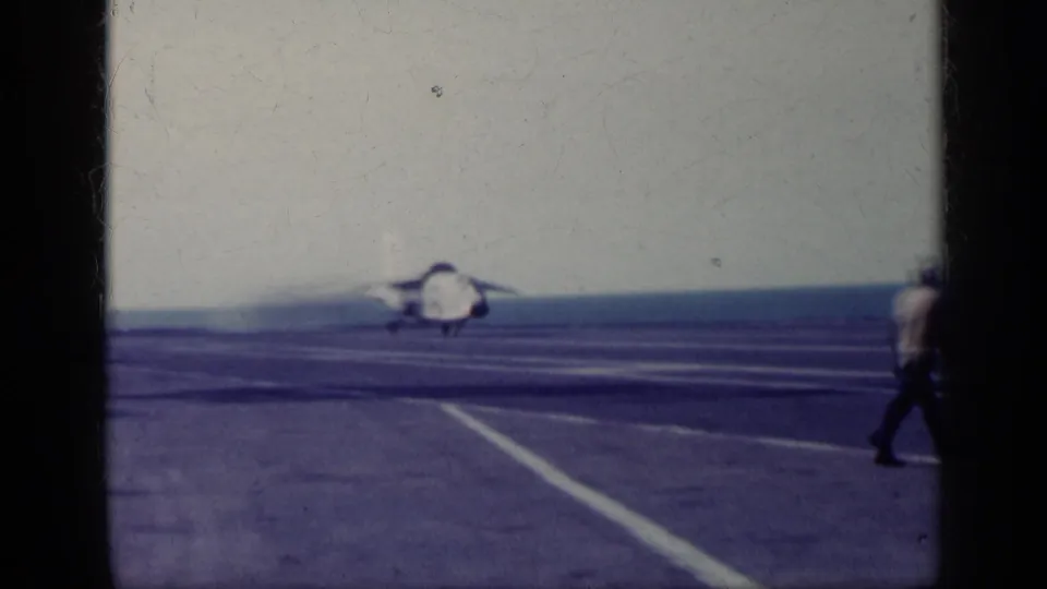 1962 Cuba-linked 8mm frame with a jet receding along the carrier deck, ocean horizon visible, and a person walking at the right edge.