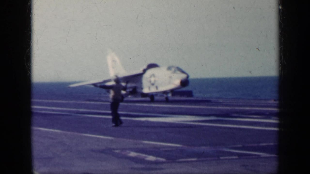 Naval jet on an aircraft carrier deck with a crew member signaling in front of it.