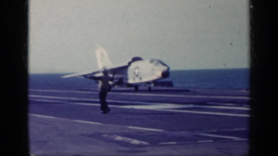 1962 Cuba-related 8mm frame showing a naval jet on a carrier deck as a deck crew member gives hand signals near the ocean horizon.