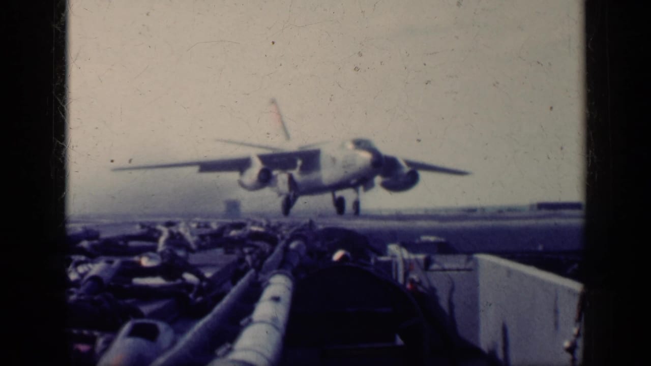 1962 Cuba-context 8mm frame showing a military jet descending toward a carrier deck, viewed past dark framing and deck equipment.