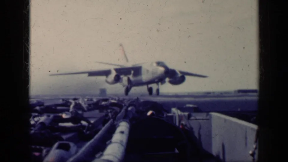 1962 Cuba-context 8mm frame showing a military jet descending toward a carrier deck, viewed past dark framing and deck equipment.