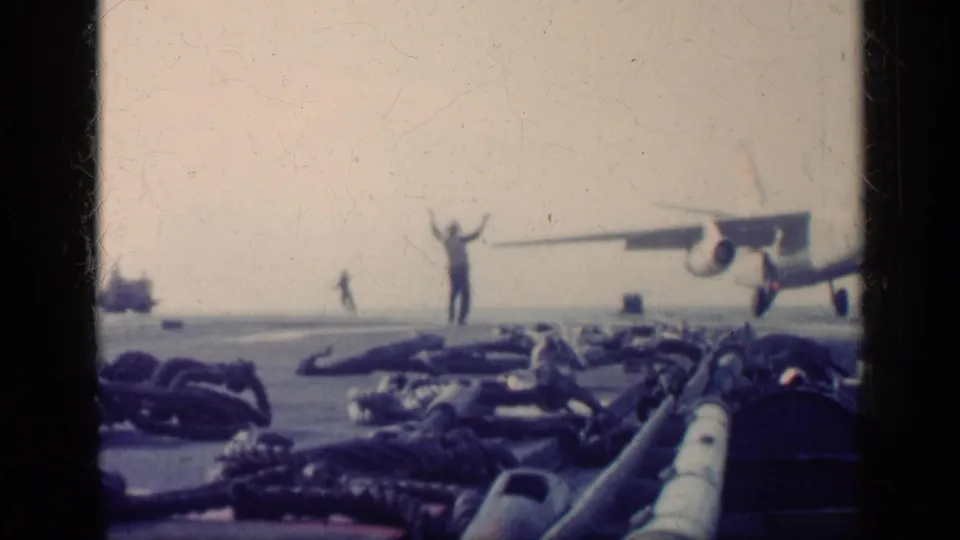 1962 Cuba-context carrier deck view with a signaler raising both arms while a military jet comes in from the right.