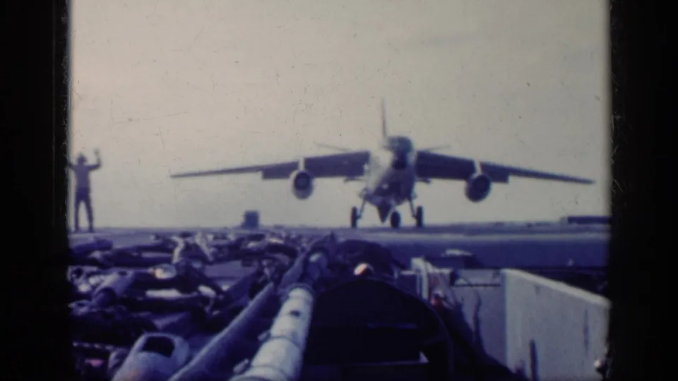 1962 Cuba-context 8mm frame showing a military jet on or just above the carrier deck, with a crew member signaling at left.