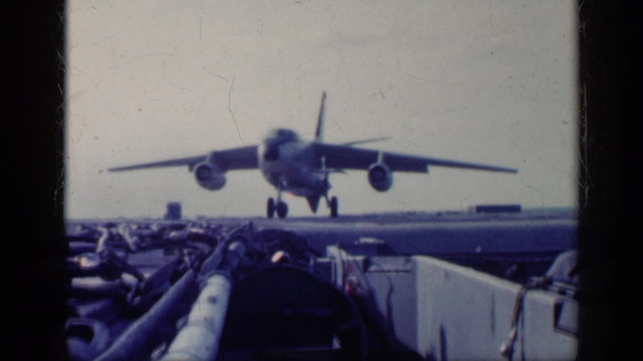 Military jet facing the camera on an aircraft carrier deck with deck equipment in the foreground.