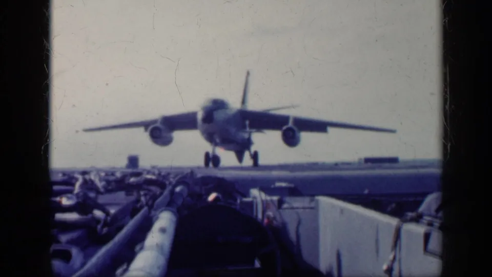 1962 Cuba-related 8mm frame showing a military jet head-on on an aircraft carrier deck, with ocean horizon and deck equipment visible.
