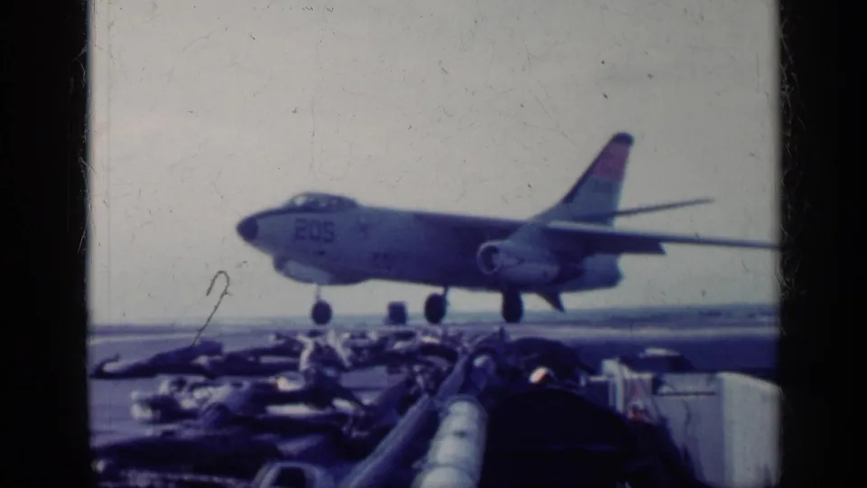 1962 Cuba-related 8mm frame showing a gray military jet marked 205 in side view on a carrier deck, with deck equipment in front.
