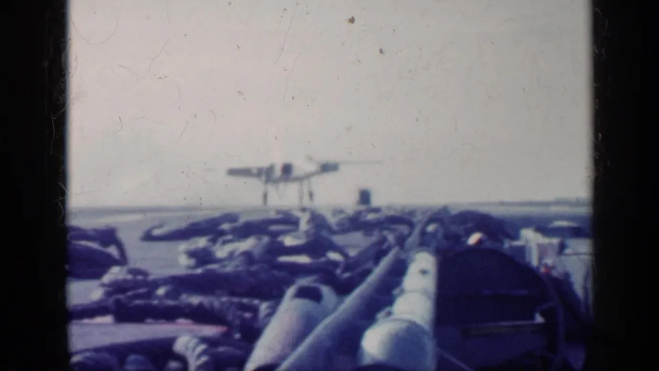 1962 Cuba-related 8mm frame with a distant military jet on the carrier deck, viewed over pipes and equipment in the foreground.