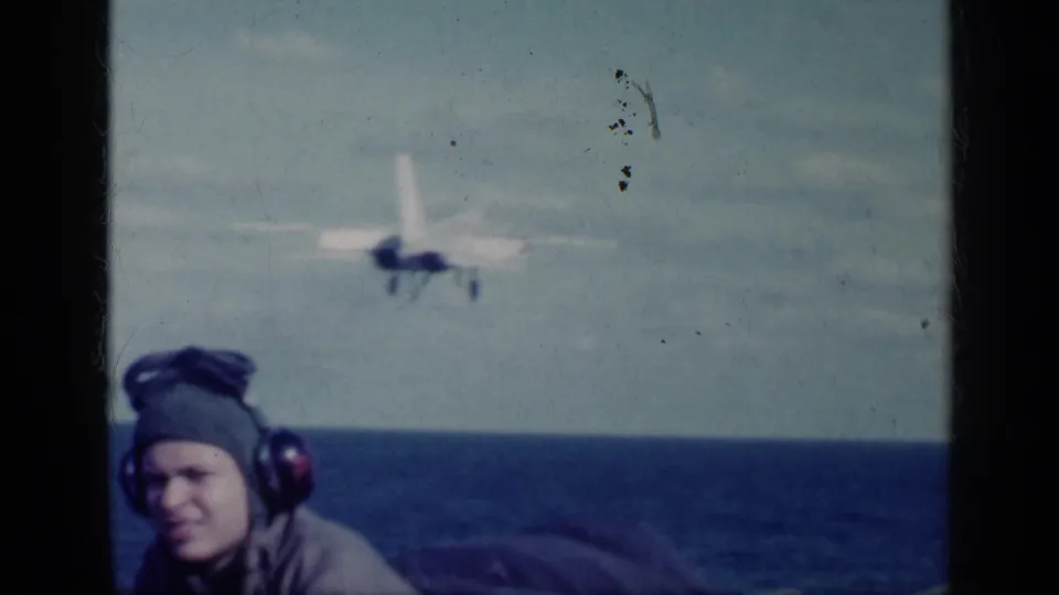 A deck observer wearing a flight helmet and headphones watches an aircraft climb over the ocean in 1962 Cuba-linked footage.